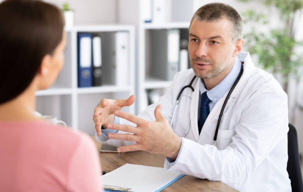 Middle aged experienced doctor talking to his patient Visiting Physician Concept. Portrait of experienced middle aged doctor talking to woman, explaining diagnosis and treatment. Therapist consulting young patient sitting at table in hospital